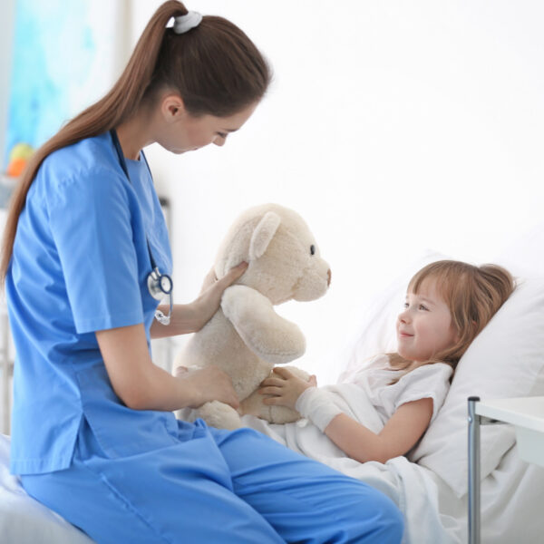 Weibliche Pflegekraft sitzt am Bett eines kranken Mädchens und zeigt ihr einen weißen Teddybären